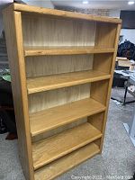 Full view of large oak bookcase with four shelves, wood grain visible, some items and furniture visible in background.