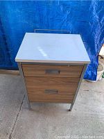 Front view of metal end table showing white top and wood finish drawers.