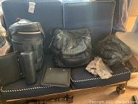 Photo shows a collection of black leather motorcycle bags, including cylindrical and rectangular shapes, along with gloves and a black portfolio holder on a blue cushioned bench.
