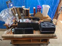 Photo of multiple office supplies, books, cordless phones, bags of pens and crayons, protractor, plastic trash can, keyboard, mouse, a car console, and poker table cover in a cardboard tube arranged on wooden shelving