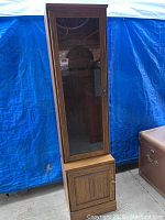Front view of the shooters cabinet against blue tarp background showing tall narrow wooden cabinet with glass door above and closed lower wood door.