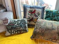 Photo showing four assorted throw pillows including a rectangular pillow with patio scene, a birds and flowers embroidered square pillow, a "Home Sweet Home" pillow, and a green botanical pillow.