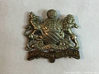 Front view of an antique WWI Manchester Regiment metal cap badge showing detailed heraldic design including a lion, unicorn, crown, motto scrolls, and 'MANCHESTER' inscription.