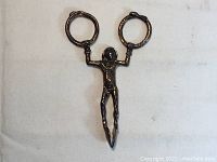 Full view of vintage metal tongs shaped like a harlequin figure with circular handles and pointed grip ends.