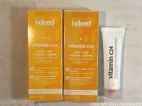 Front view of two orange boxes of Indeed Vitamin C24 cream and one white tube with orange tip of the cream visible on a white background.