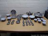 Large display of Asian porcelain dishware laid out on table including bowls, spoons, teapot, tea cups, and plates all featuring floral patterns mainly in black, pink, and green