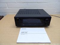 Front angle view of black Denon AVR-1707 AV surround receiver sitting on wood table with user manual placed in front.