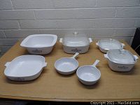 Seven white ceramic Corning Ware dishes with vintage blue floral Cornflower pattern, assorted shapes and sizes, some with glass lids, on a wood table against a cinderblock wall