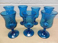 Front view of seven handmade blue glass drinking glasses with swirl design, displayed on a light surface against a plain white brick background.