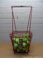 Red metal tennis ball pickup rack filled with green tennis balls seen from the front against a white brick wall.