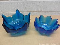 Two unique blue glass dishes side by side on a wooden surface against a white brick background. The left dish is larger with pointed petal edges; the right is smaller and more rounded.