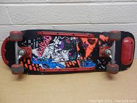 Top view of skateboard showing colorful graphics under deck and red wheels, also black deck with red ends