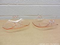 Pair of vintage pink glass dishes with bow-shaped handles, shown side by side on a flat surface against a gray textured wall.