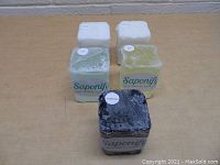 Five soap making blocks packed in original plastic wrap under Saponify brand. Varied colors white, green, yellow, and black charcoal.