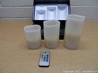 Three off-white battery operated flameless candles in front of a black box and a remote control on a wooden surface.
