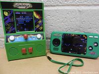 Two Galaga mini arcade games side by side against a white textured wall. Left is a mini arcade cabinet with joystick, buttons, speaker grills, and top marquee art. Right is a green handheld arcade with screen, directional pad, A and B buttons, and wrist strap.