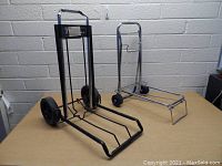 Two folding carts on a wooden surface against a brick wall. One black cart has larger wheels, the other silver cart has smaller wheels and a foldable hook.