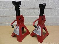 Pair of red 2 ton capacity metal jack stands with triangular bases and black adjustable ratcheting supports, side by side on a table in front of a brick wall.