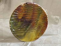 Photo of the front side of the round slag glass plate showing marbled coloration yellow, brown, green, and reddish hues with wavy texture.