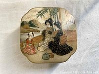 Top view of small hand-painted Japanese porcelain box lid showing traditional scene with figures and outdoor setting.