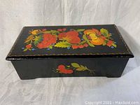 Large black lacquer rectangular box with colorful floral painting on lid and side, placed closed on white fabric background.