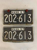 Front of two 1957 Massachusetts license plates numbered 202-613. One plate has two red reflectors attached.