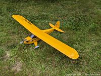 Yellow balsa wood Piper Cub RC model airplane placed on grass, showing front angle with visible motor and propeller.