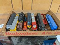 Overview in cardboard box showing silver streamlined loco, yellow coach, black diesel, turquoise maintenance car, small orange and red automobiles, red gondola