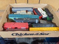Box of mixed HO gauge train cars and engines showing variety of freight cars including tanker, boxcars, flatcar with crates.