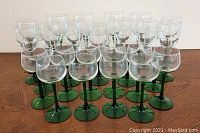 Front view of 28 clear stemware glasses with green stems and bases arranged in rows on wooden surface