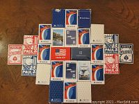 Top-down view of twenty-seven packs of playing cards spread out on a wooden surface. Visible brands include Carnival, Royal Caribbean, Delta Airlines, and Paulson Casino. Some card boxes show signs of use and damage.