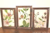 Three framed hand-painted glass art pieces placed side by side showing botanical floral designs with green leaves in wood-tone frames.