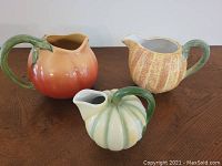 Three fruit-shaped ceramic pitchers together on a wooden surface showing size and color variations
