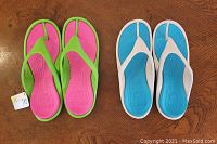 Two pairs of Crocs flip-flop sandals on a wooden surface. The left pair is lime green with fuchsia footbed and a visible size tag. The right pair is light gray with white straps and a teal footbed.