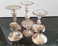 Front view of six silver or silver plated candlesticks showing various styles and sizes on black surface.
