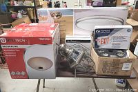Photo shows various boxed lighting fixtures grouped on a table including a Utilitech ventilation fan with light, Nutone outdoor wall sconce, and a Lithonia Lighting exit sign.