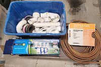 Plastic bin with assorted white PVC plumbing fittings and a copper refrigeration coil coiled on the ground nearby, alongside a boxed misting system.