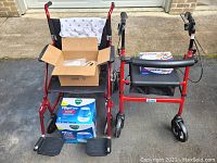Photo showing red collapsible transport chair with black seat and footrests, box of foley supplies on seat, Vicks humidifier box on ground, and red rolling walker with padded seat and storage pouch nearby