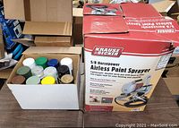 Boxed Krause & Becker airless paint sprayer with spray gun and hose on table next to a box of spray paint cans.