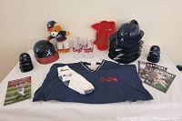 Full contents of the Braves souvenir package laid out on white surface. Includes jacket, helmets, mugs, teddy bear, foam hand, and game-day programs.