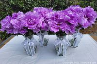 Four large glass vases filled with clear glass decorative rocks and large artificial lavender peonies, displayed on a white cloth outdoors.