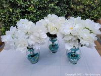 Three large glass vases about 11 inches tall, each filled with multiple large white artificial peony flowers and colorful blue and amber decorative glass rocks at the base inside the vase, set outdoors on a white cloth.