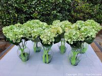 Front view of six large glass vases with artificial green hydrangeas and decorative glass rocks on a white table.