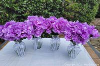 Four clear glass vases filled with artificial lavender peony flowers and clear decorative glass rocks on white tablecloth outdoors.
