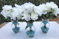 Three glass vases with white artificial peonies and blue glass rocks on a table outside with greenery background.