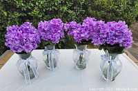 Four artificial lavender hydrangea flower arrangements displayed on a white tablecloth with green hedge background. Each arrangement is in a clear glass vase filled with clear decorative rocks and tied with white ribbon bows.
