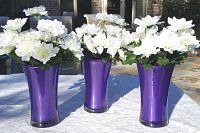Set of three purple hourglass-shaped vases with artificial white chrysanthemum flowers arranged inside, placed outdoors on a white textured tablecloth.
