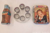 Top-down photo showing six Coca-Cola lowball glasses in a round arrangement along with the Coca-Cola bottle-shaped metal tin on the left and rectangular metal tray on the right