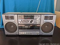 Front view of Hitachi TRK-9011HC boombox showing detachable speakers, cassette player, tuning and volume knobs, and graphic equalizer sliders.