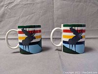 Front view showing two ceramic mugs with a moose silhouette and multicolor stripe top rim.